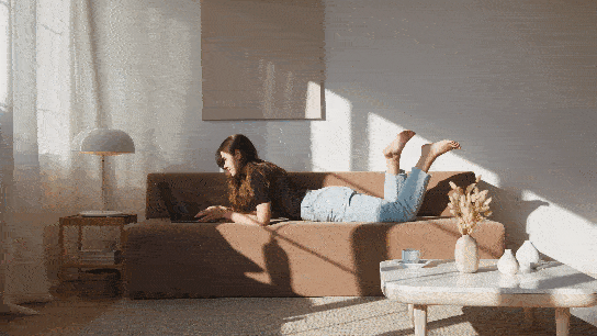 A beautiful girl lying comfortably on her sofa and wearing a black blouse and jeans, barefoot typing on her Laptop.