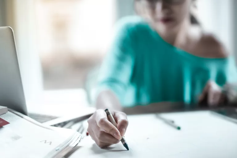 A beautiful girl in a green blouse is drawing on paper with colored markers in front of her desk.