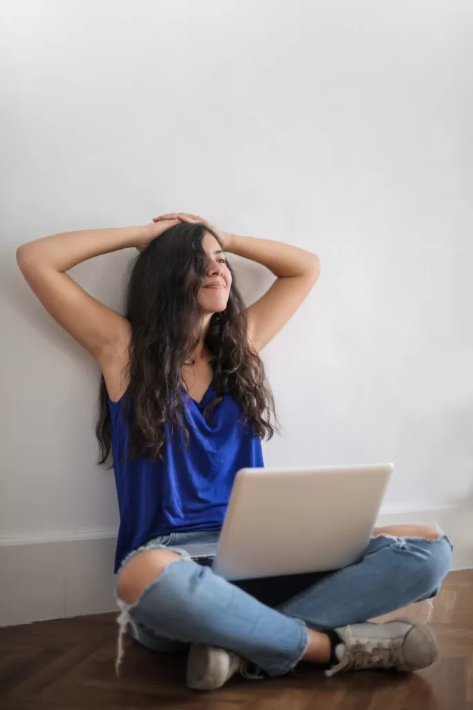 Happy girl sitting on the floor wearing blue jin and blue blouse, happy with hands behind her head, with a laptop in her legs.